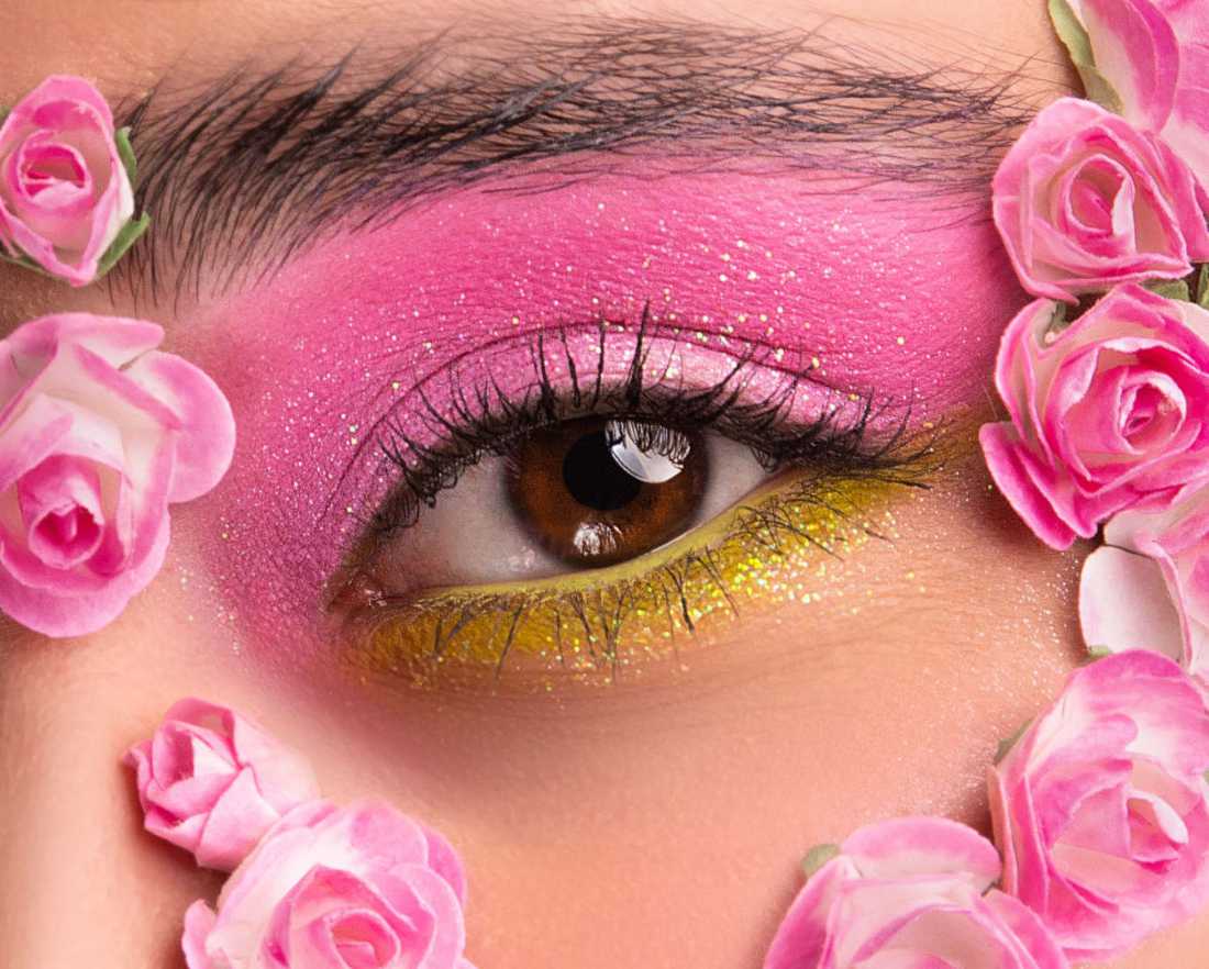 Fotografía close-up de un ojo color café con maquillaje rosa y verde y flores a su alrededor como adorno.