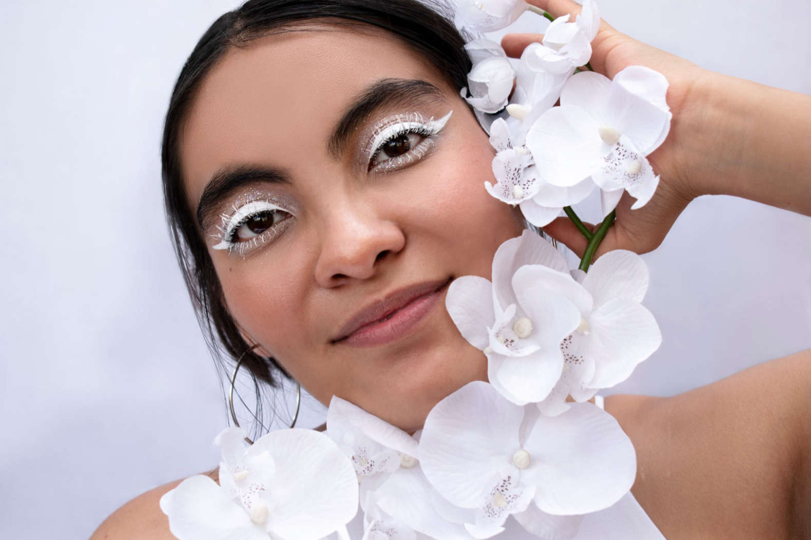 Fotografía close-up de una joven con maquillaje y atuendo blanco sosteniendo un racimo de orquídeas.